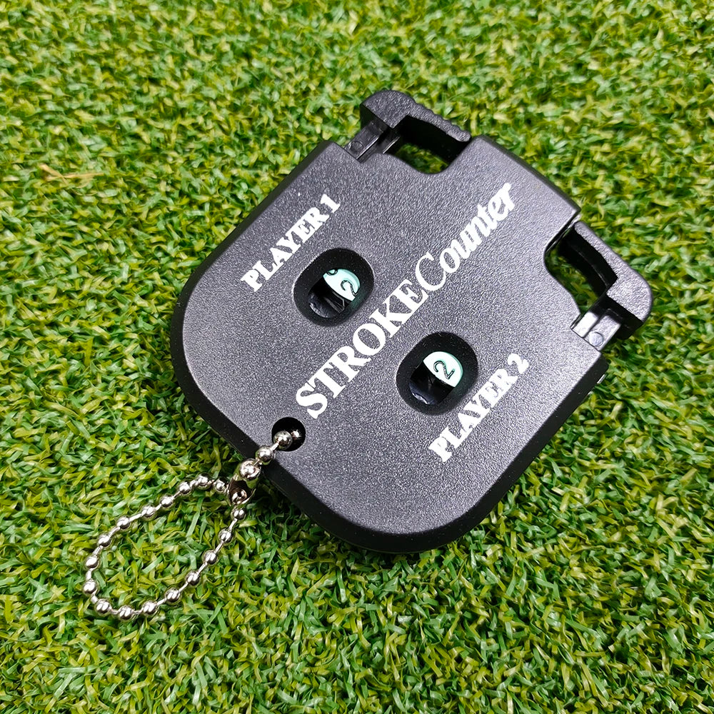 A black portable golf score counter with a chain attached, lying on green grass.