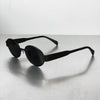 A pair of classic oval metal sunglasses in black resting on a light gray surface.