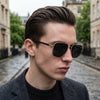 A close-up portrait of a man wearing classic square aviator sunglasses with gold accents.