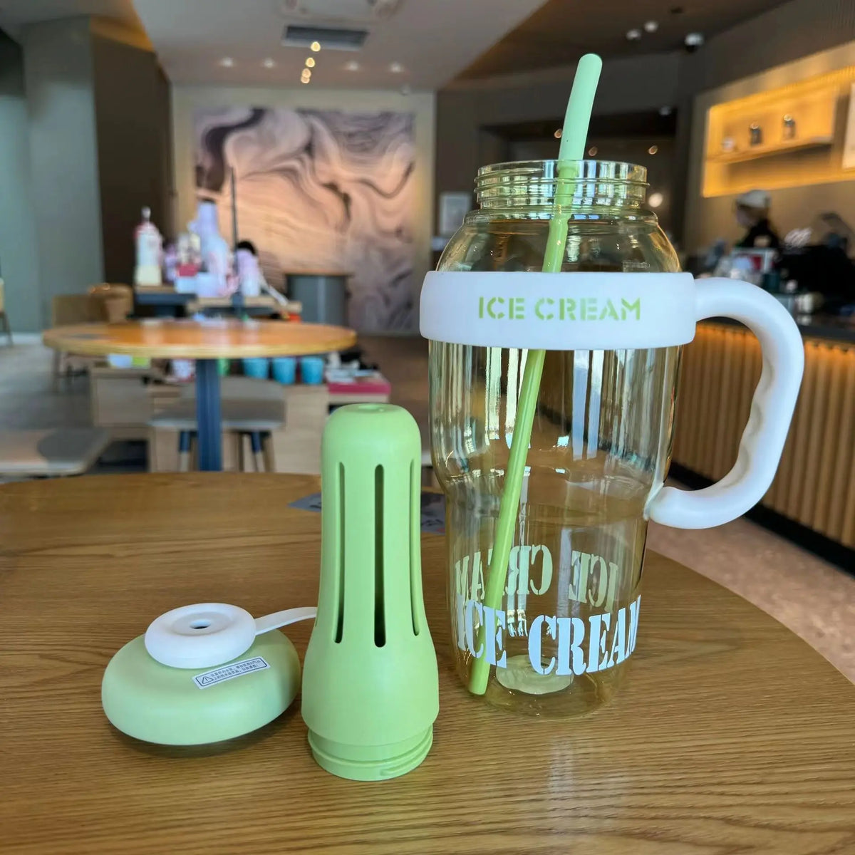 A clear glass water bottle with a handle and straw, labeled ICE CREAM, sits on a wooden table next to a lime green silicone b