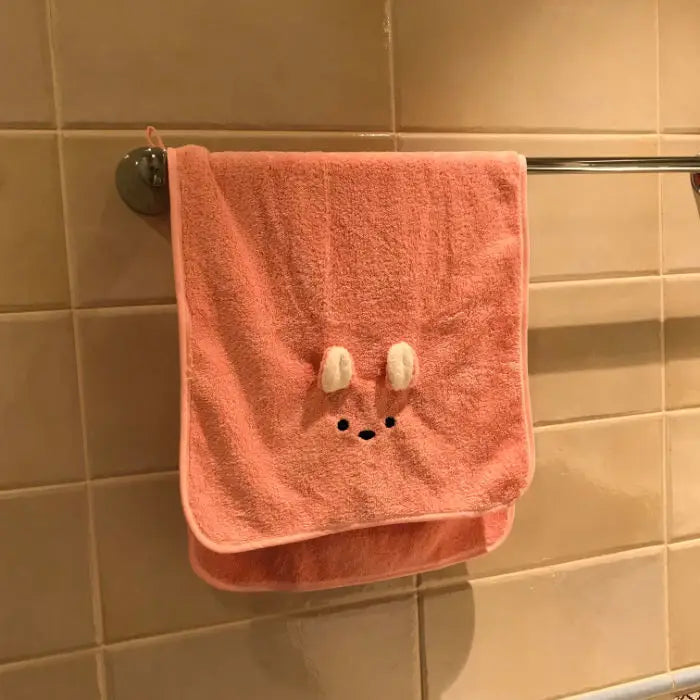 A pink coral fleece face towel with a cute bunny face design hanging on a metal rack.