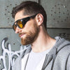 A man wearing black retro steampunk goggles sunglasses with amber lenses and a beard, looking to the side.