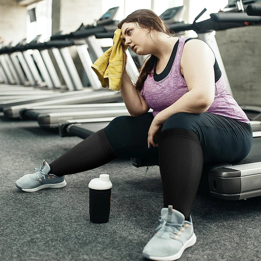 A woman wearing compression socks is sitting on a gym floor with various exercise equipment in the background.