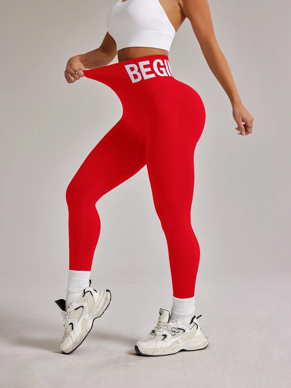 Eine Frau in roten, hoch taillierten, nahtlosen Yoga-Leggings und einem weißen Crop-Top steht auf einem hellgrauen Boden.
