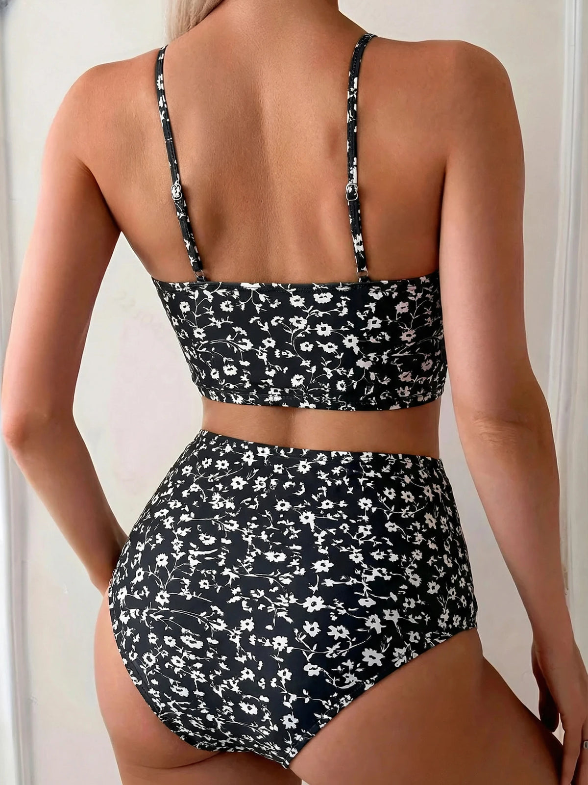 Una mujer modela un bikini de dos piezas con estampado floral negro de cintura alta desde la espalda.