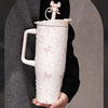 A white insulated thermos mug with pink floral inlay details is held against a dark gray fabric.