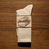 A white tube sock with a brown waistband featuring the word Quality printed on it.