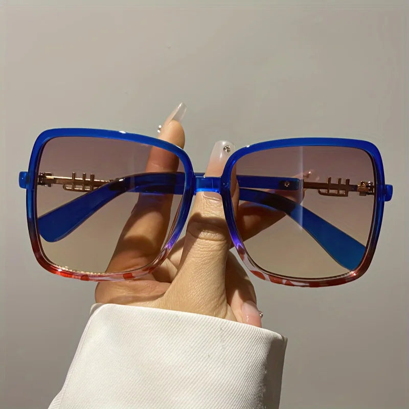 A hand holds a pair of retro two-tone gradient sunglasses with blue and amber lenses against a light background.