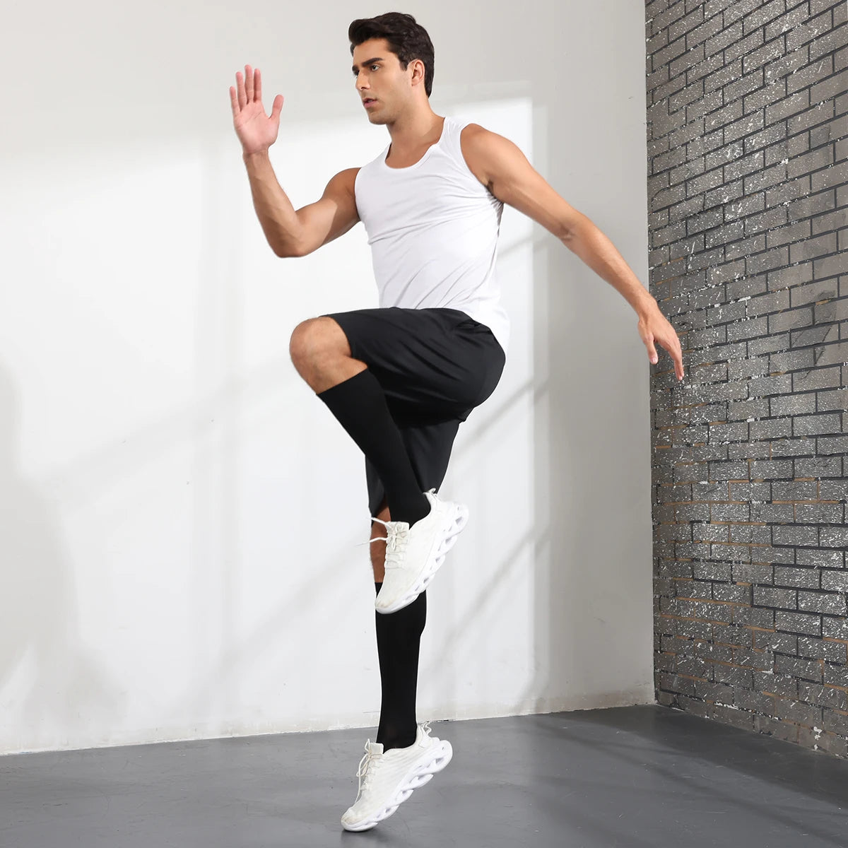 A young man wearing white tank top and black athletic shorts is demonstrating a gesture while standing in front of a brick wa