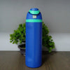 A blue insulated stainless steel water bottle with a teal cap and dual drinking spout, set against a green plant background.