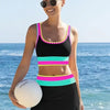 A woman wearing a black swimsuit with neon pink and teal color blocks stands near the ocean.