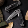 A black multi-functional folding survival knife is shown with a sheath and a leaf on a dark background.