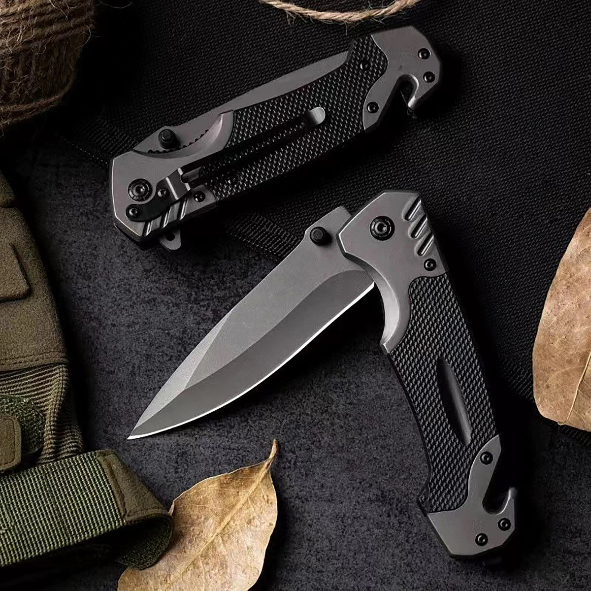 A black multi-functional folding survival knife is shown with a sheath and a leaf on a dark background.