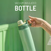 A hand holds a light green vacuum insulated bottle with a silver cap against a blurred green background.